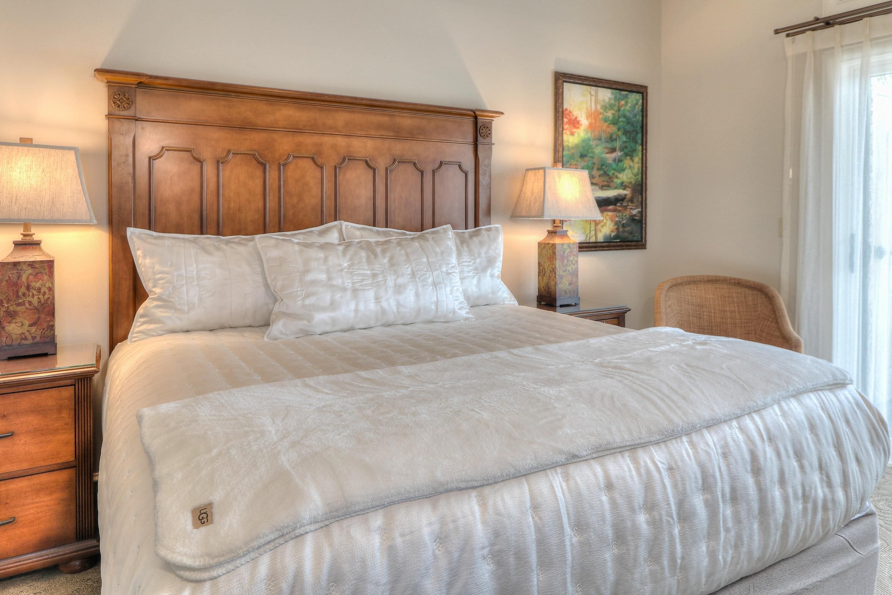 A neatly made bed with a wooden headboard, two bedside lamps, a chair, and a painting on the wall.