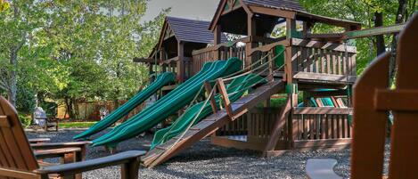 A wooden play structure with slides and a small house, surrounded by trees and gravel.