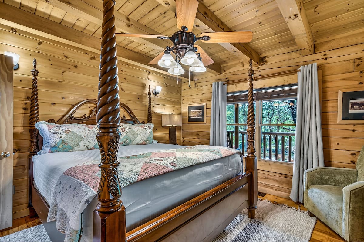 A four-poster bed with a quilt, a wooden ceiling fan, and a window with curtains.