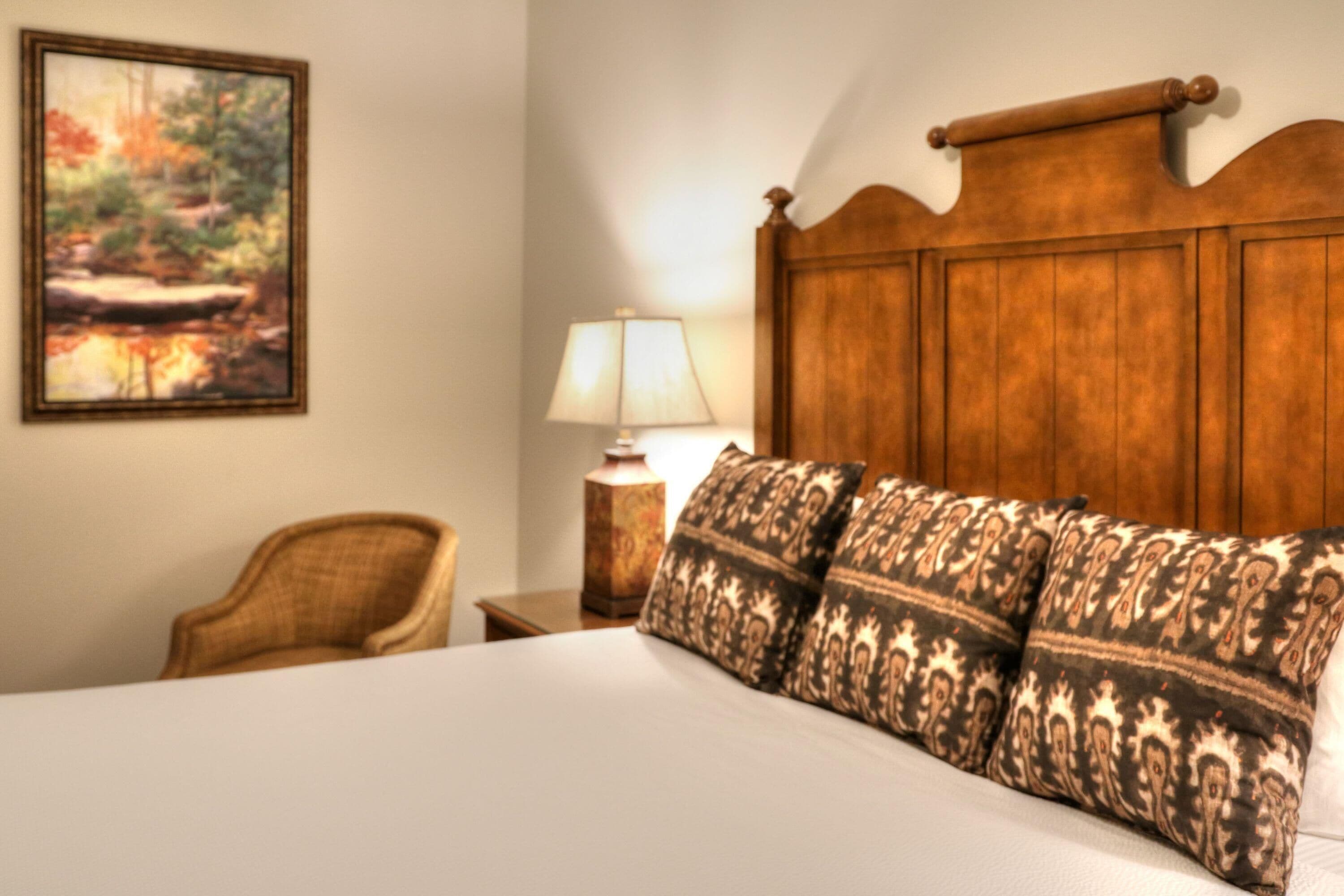 Una habitación de hotel con cabecera de madera, cama con almohadas estampadas, una silla, una mesita de noche con lámpara y un cuadro en la pared.