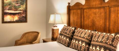 A hotel room with a wooden headboard, a bed with patterned pillows, a chair, a bedside table with a lamp, and a painting on the wall.