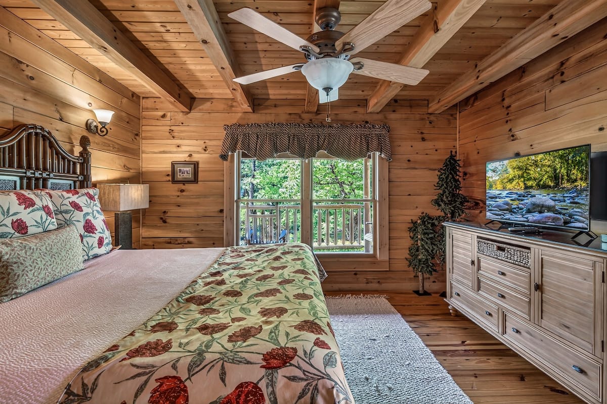 A wooden cabin bedroom with a bed, a ceiling fan, a TV, and a window with a view of trees.
