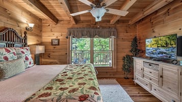 A wooden cabin bedroom with a bed, a ceiling fan, a TV, and a window with a view of trees.