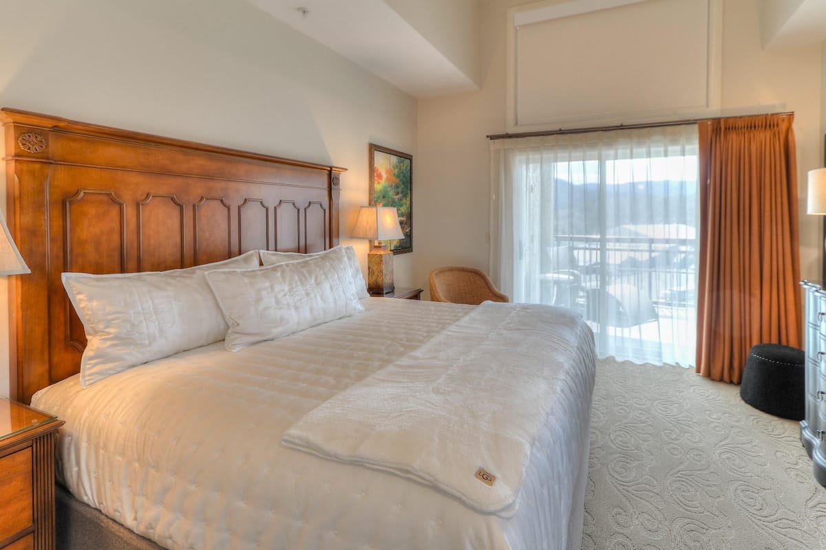 A hotel room with a large bed, a wooden headboard, a nightstand with a lamp, a chair, and a view of the outdoors through a window with curtains.