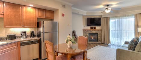A modern kitchen with wooden cabinets, a stainless steel refrigerator, and a dishwasher. There is a dining table with chairs and a vase of flowers. A fireplace is visible in the background, and a ceiling fan is above the dining area.