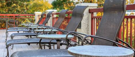 Outdoor seating with metal chairs and round tables, a red railing, and trees in the background.