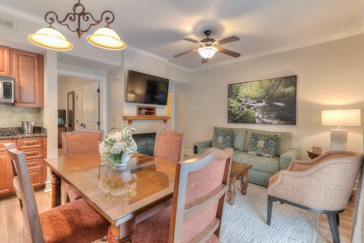 A dining area with a wooden table and chairs, a ceiling fan, and a TV in the background.