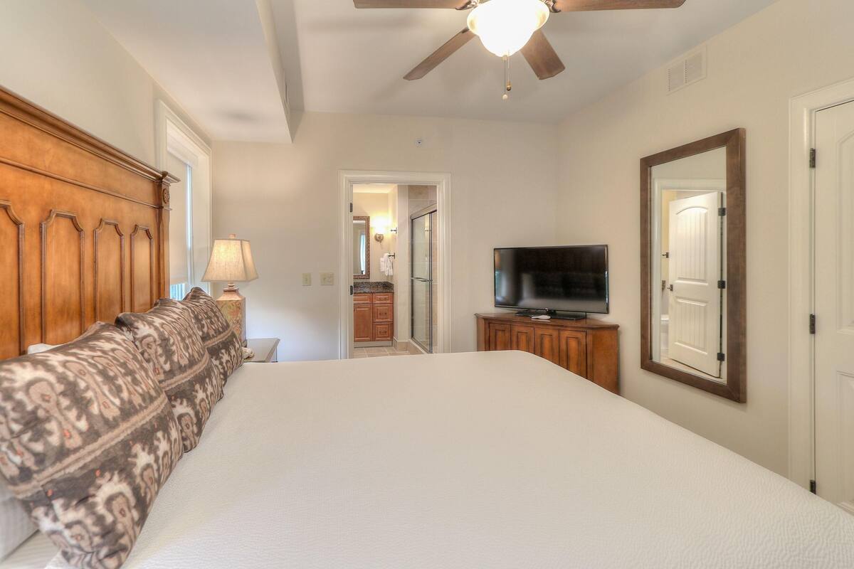 A bedroom with a large bed, a ceiling fan, a television, and a wooden headboard.