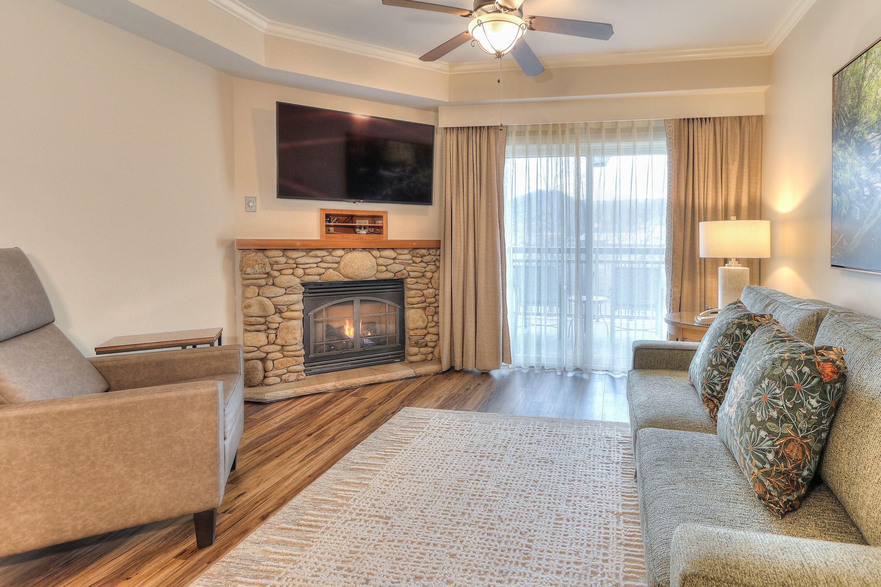 A living room with a stone fireplace, a flat-screen TV mounted above it, a ceiling fan, and a large window with curtains.