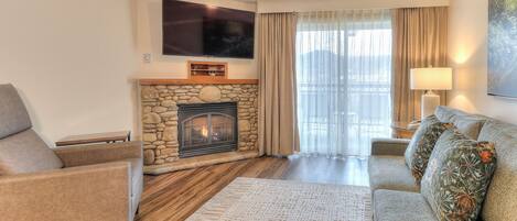 A living room with a stone fireplace, a flat-screen TV mounted above it, a ceiling fan, and a large window with curtains.