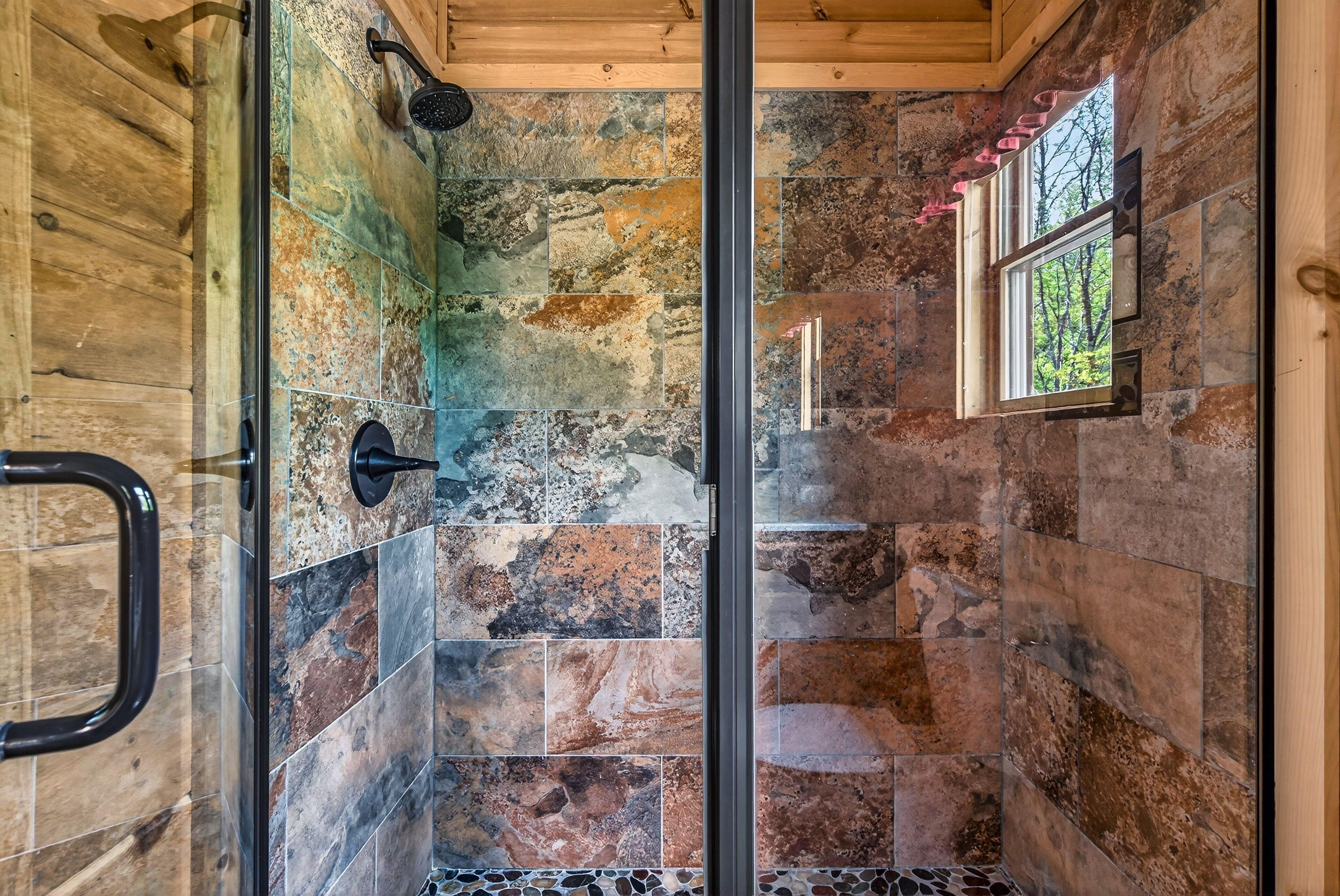Ducha con puerta de vidrio, baldosas de piedra y ventana con vista a árboles.