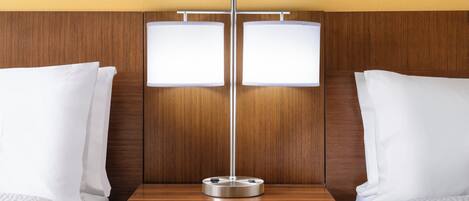 Una habitación de hotel con cabecera de madera, dos camas con ropa blanca y una mesita de noche con lámpara.