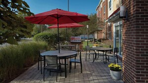 An outdoor patio with round tables and chairs, red umbrellas, and a brick building in the background. - TownePlace Suites by Marriott Winchester (Winchester)
