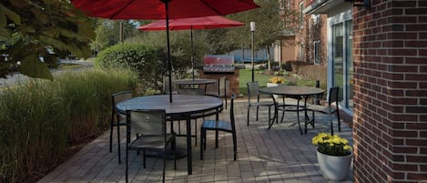 Un patio al aire libre con mesas y sillas redondas, sombrillas rojas y un edificio de ladrillo al fondo.