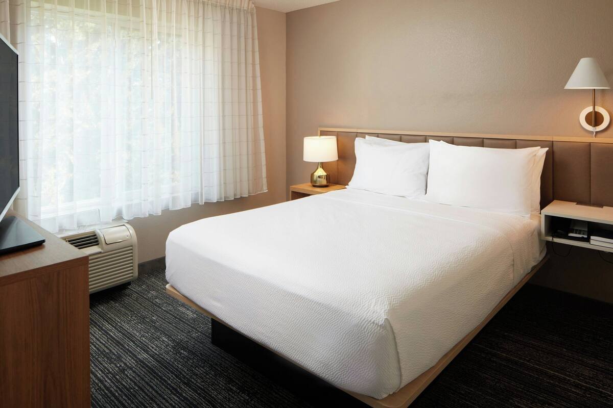 Hotelkamer met een groot bed, nachtkastje met lamp, televisie op een bureau en raam met dunne gordijnen.