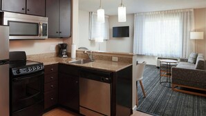 A compact kitchen with a microwave, stove, dishwasher, and sink. There is a dining area with a table and chairs, and a living room with a sofa. - TownePlace Suites by Marriott Winchester (Winchester)