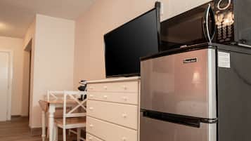 A compact kitchen with a stainless steel refrigerator, a microwave, and a flat-screen TV mounted above it. There is a wooden dining table with chairs and a white cabinet with drawers.