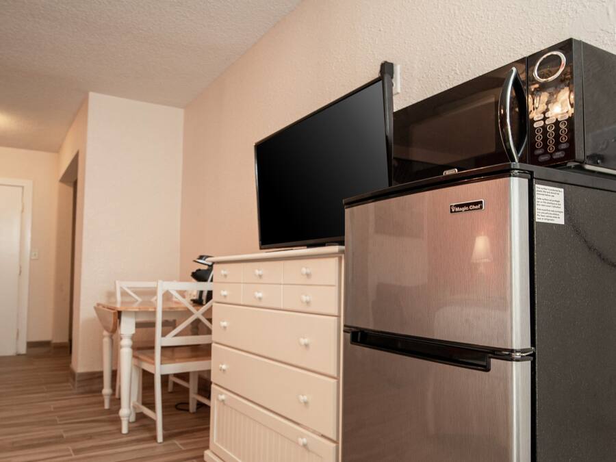 A compact kitchen with a stainless steel refrigerator, a microwave, and a flat-screen TV mounted above it. There is a wooden dining table with chairs and a white cabinet with drawers.