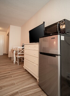 Cocina compacta con refrigerador de acero inoxidable, microondas y televisor de pantalla plana montado encima. Cuenta con una mesa de comedor de madera con sillas y un armario blanco con cajones.