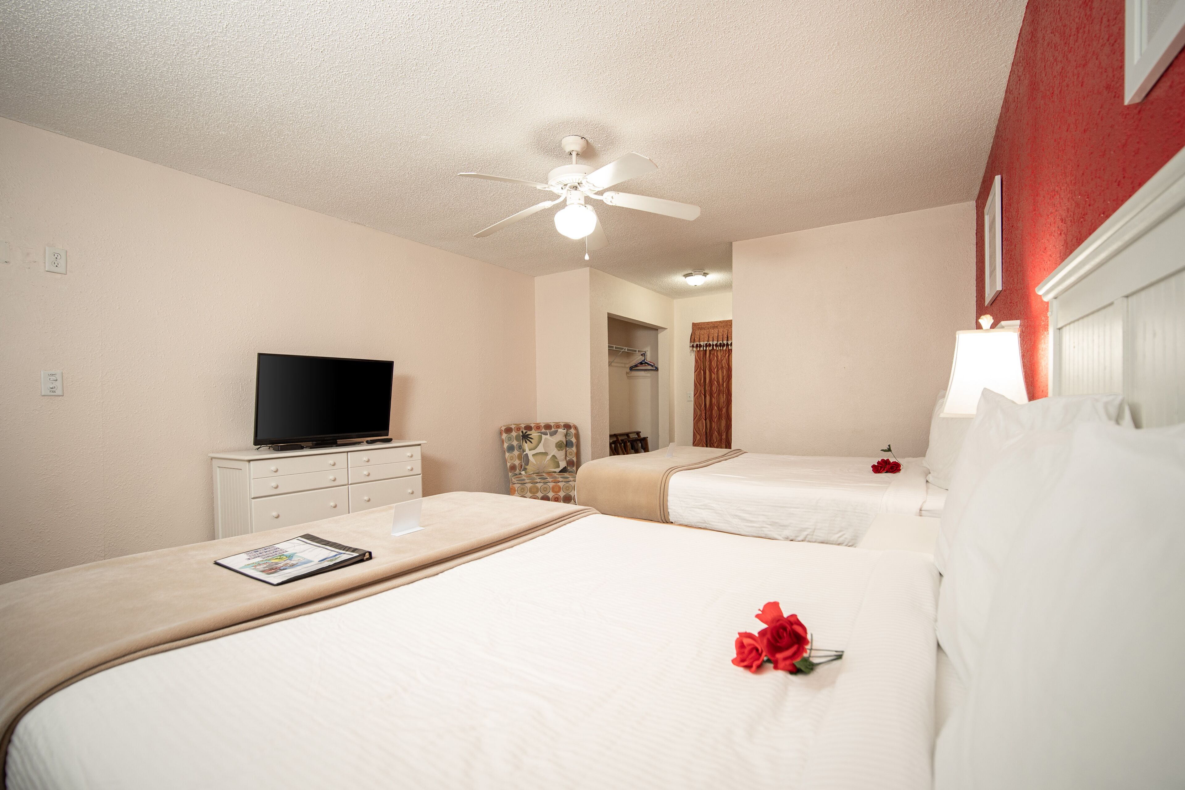 Quarto de hotel com duas camas, um ventilador de teto, uma televisão sobre uma cômoda e uma rosa vermelha em uma das camas.