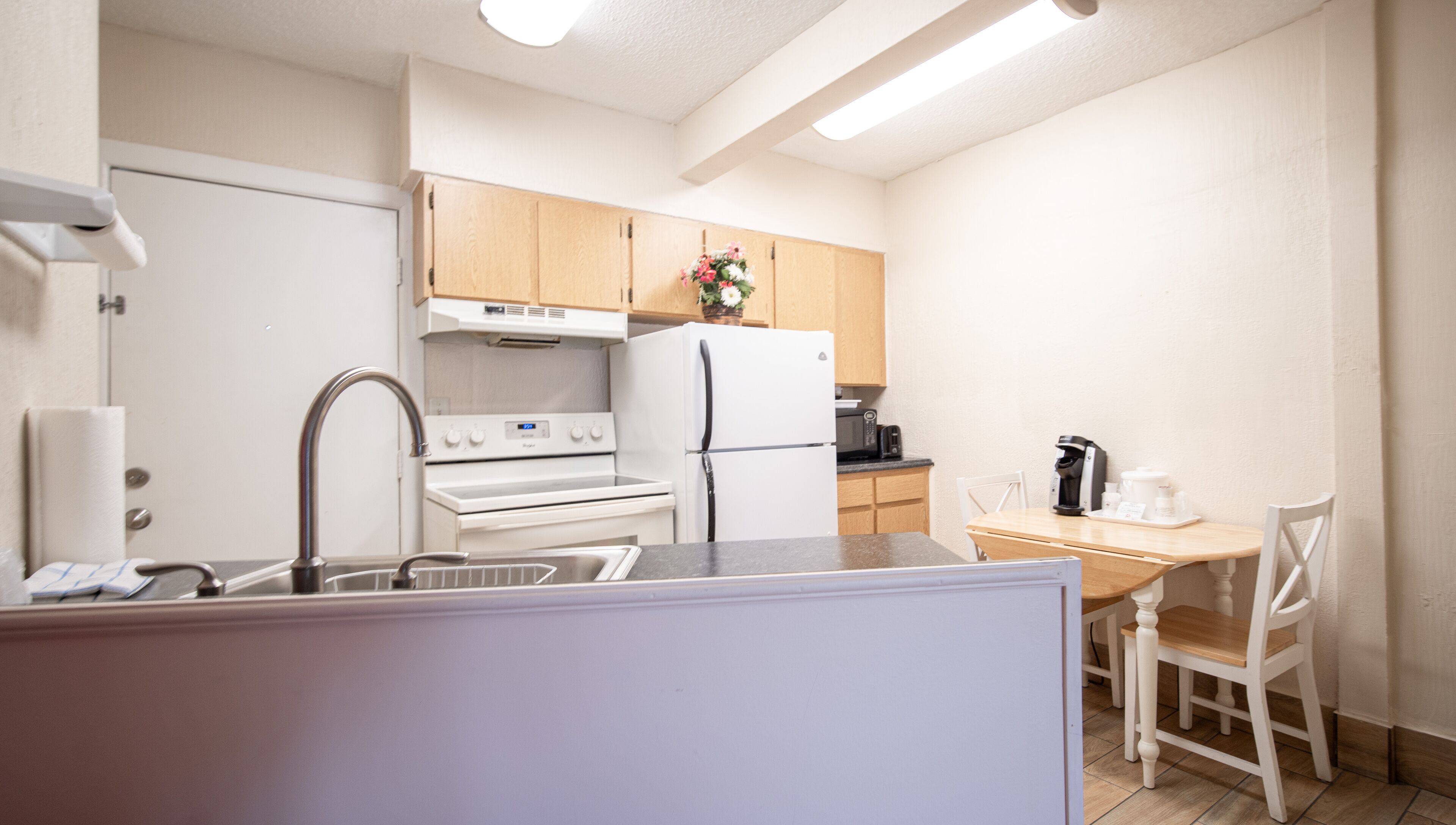 A compact kitchen with white appliances, a wooden dining table, and a single white chair.