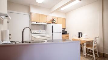 A compact kitchen with white appliances, a wooden dining table, and a single white chair.