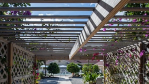 Een overdekte gang met houten constructie en lattenpanelen, die uitkomt op een tuin met planten en bloemen.