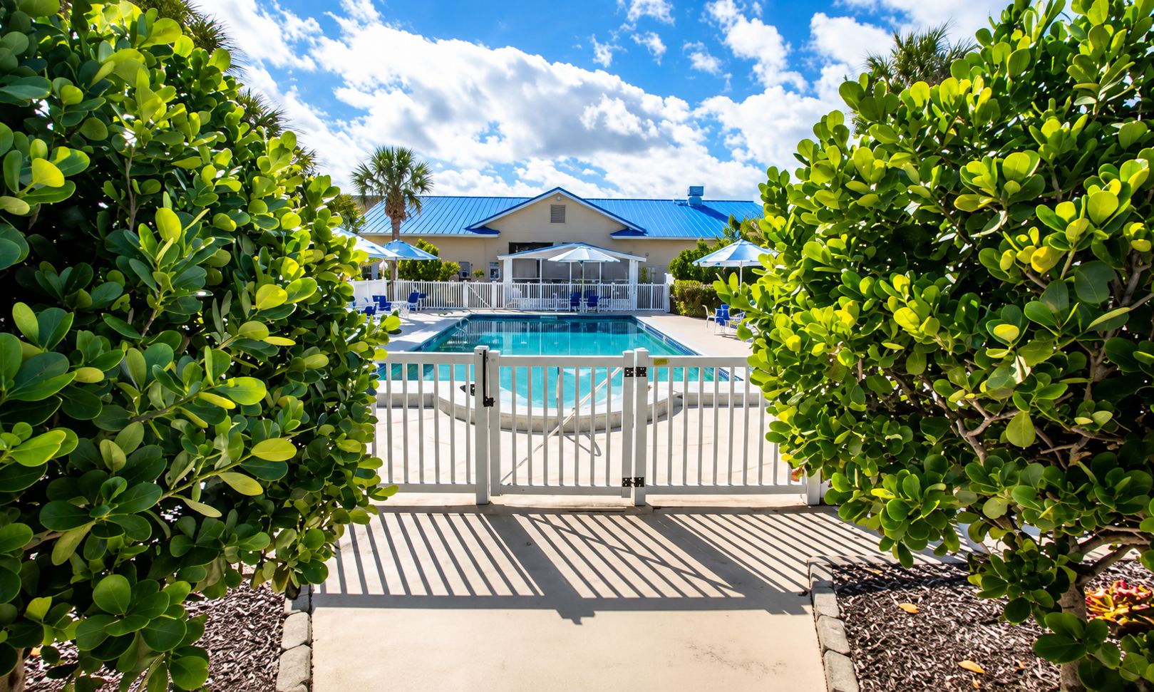 A pool area with a white fence, a covered seating area, and a building with a blue roof in the background.