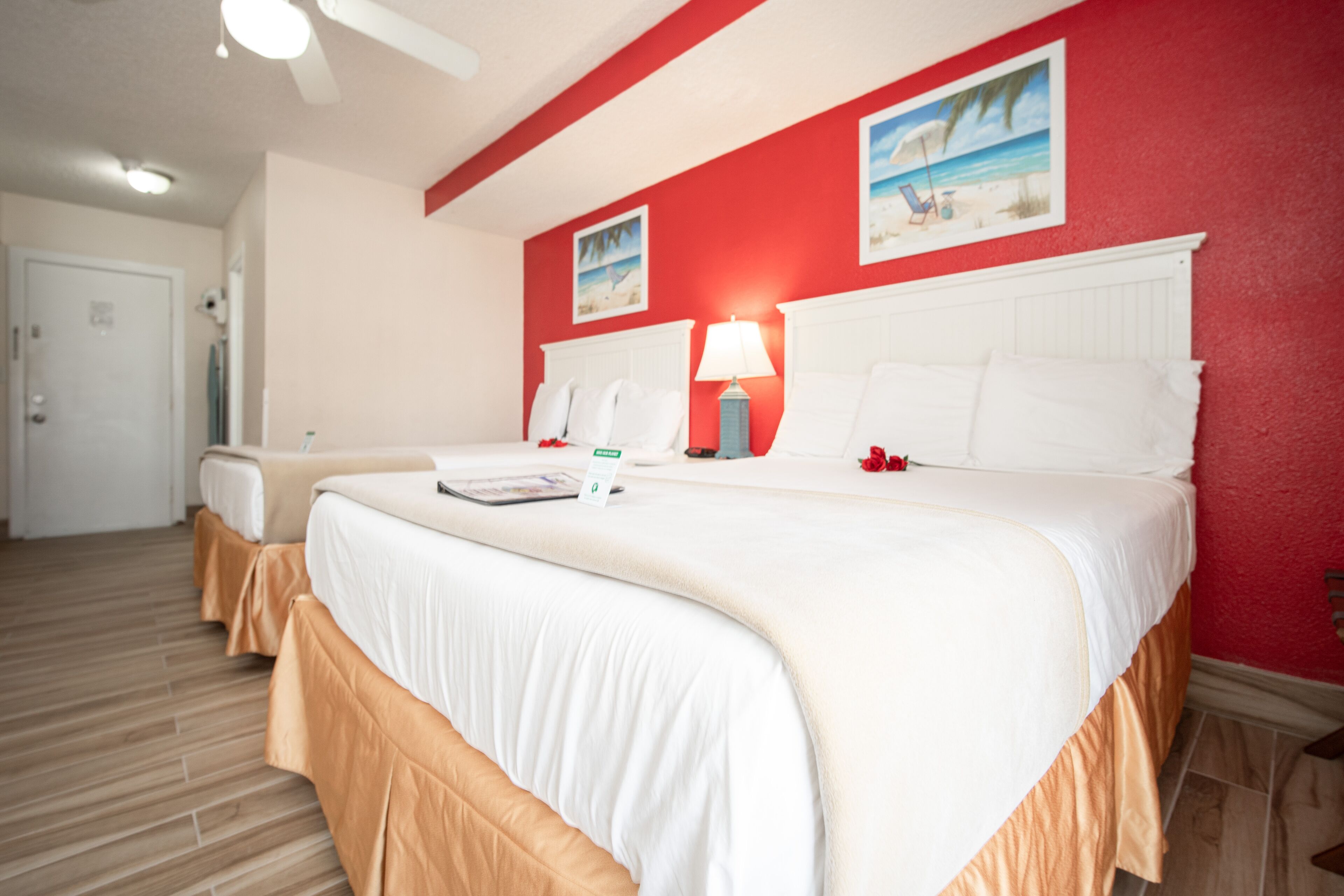 A hotel room with two beds, a red accent wall, and a beach-themed picture.