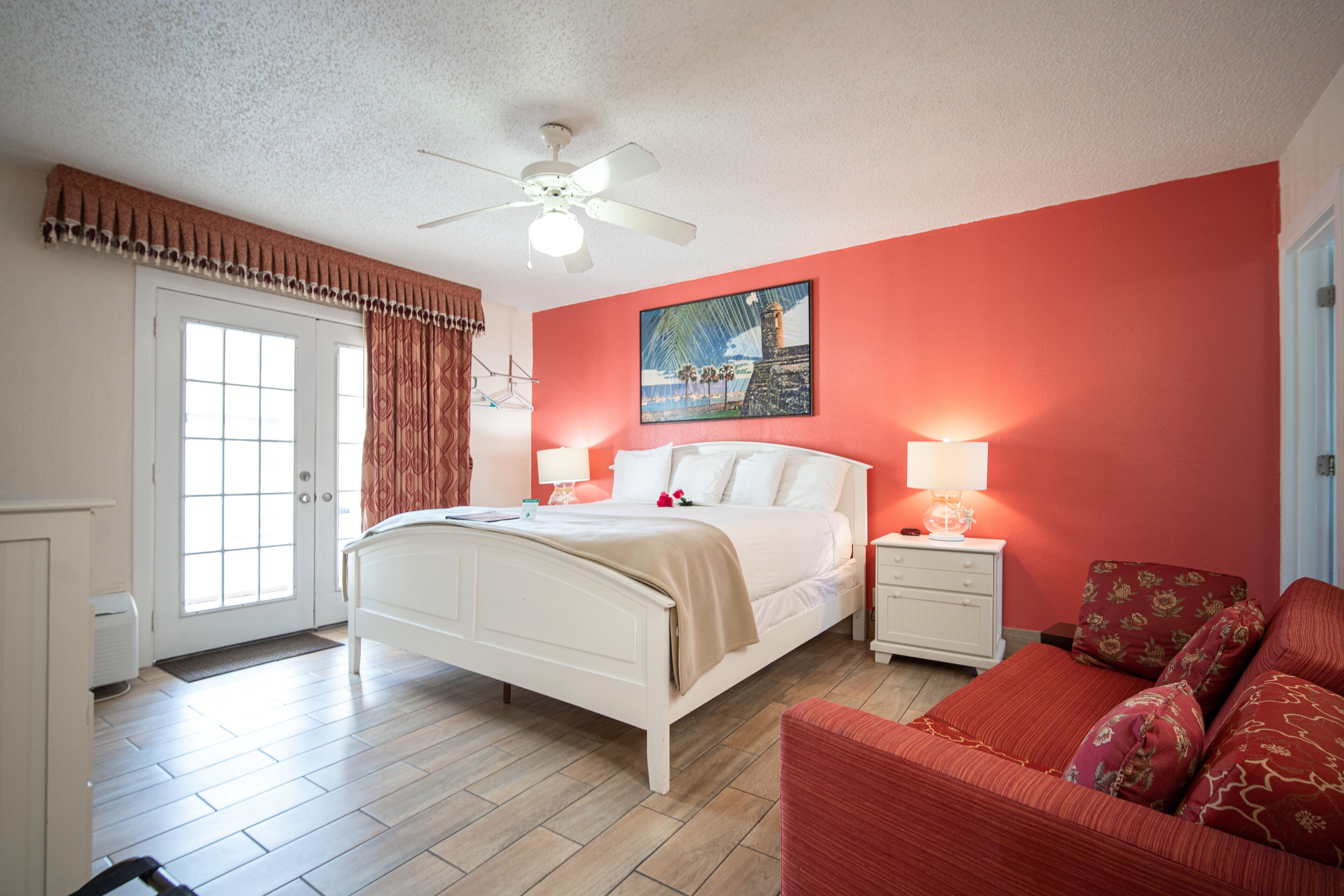 A bedroom with a bed, bedside tables, a nightstand, a red couch, a ceiling fan, a door, and a window.
