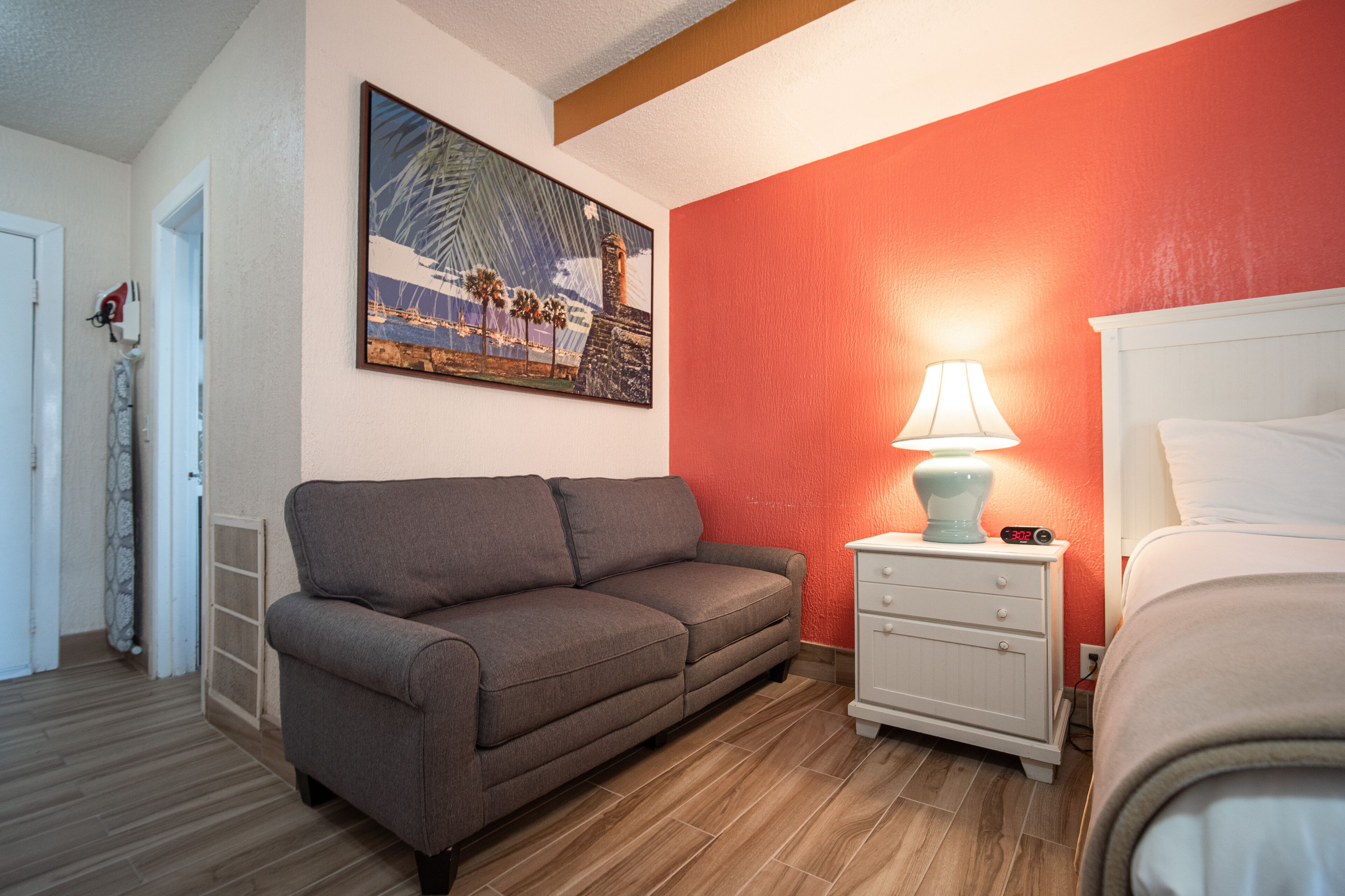 A room with a grey sofa, a white bedside table with a lamp, and a bed with white bedding.