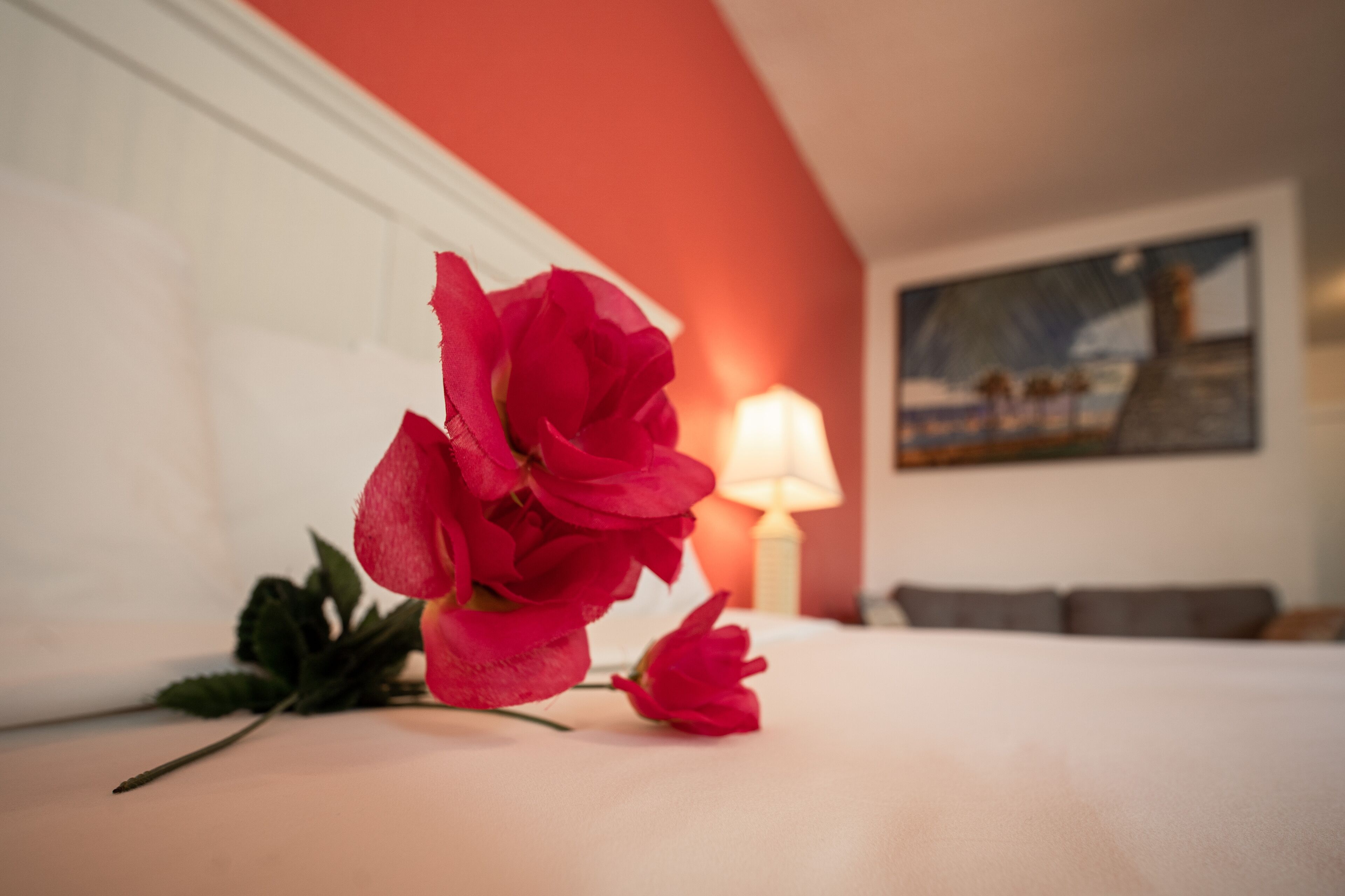 Ein Bett mit einer einzelnen roten Rose, einer Lampe und einem Gemälde an der Wand.