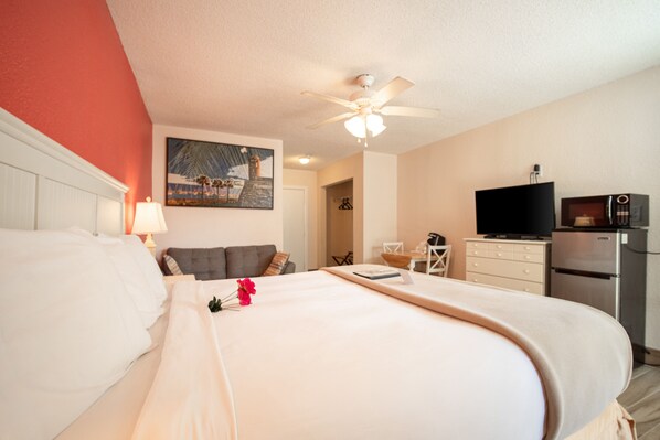 Habitación de hotel con cama, ventilador de techo, televisor y refrigerador.