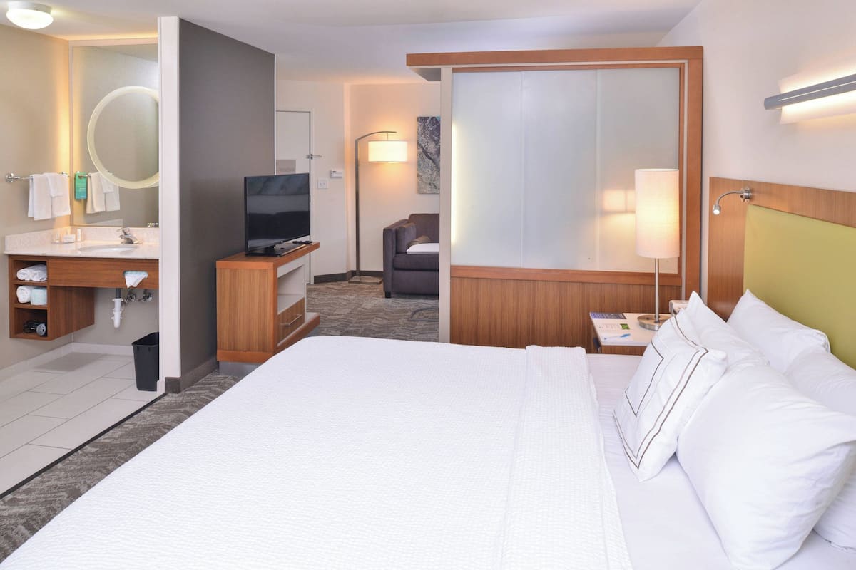 A hotel room with a large bed, a TV, a desk, a chair, a lamp, and a bathroom with a mirror and sink.