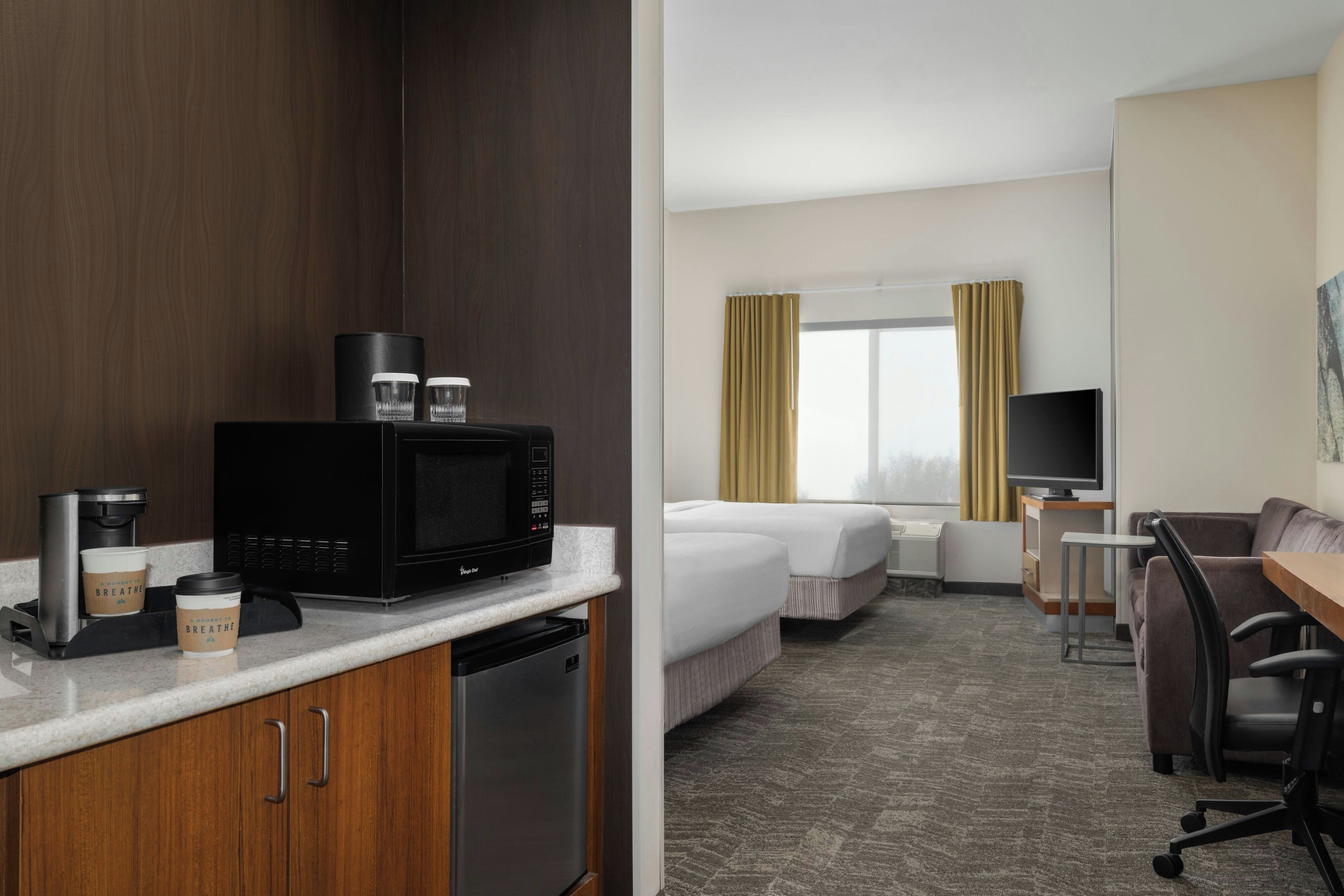 Hotelkamer met kitchenette, bed, bureau, stoel, televisie en raam met gordijnen.