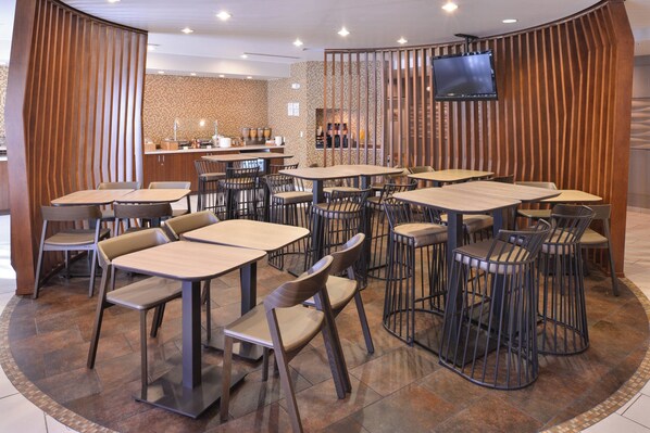 A modern restaurant with multiple tables and chairs, a kitchen area, and a TV mounted on the wall.