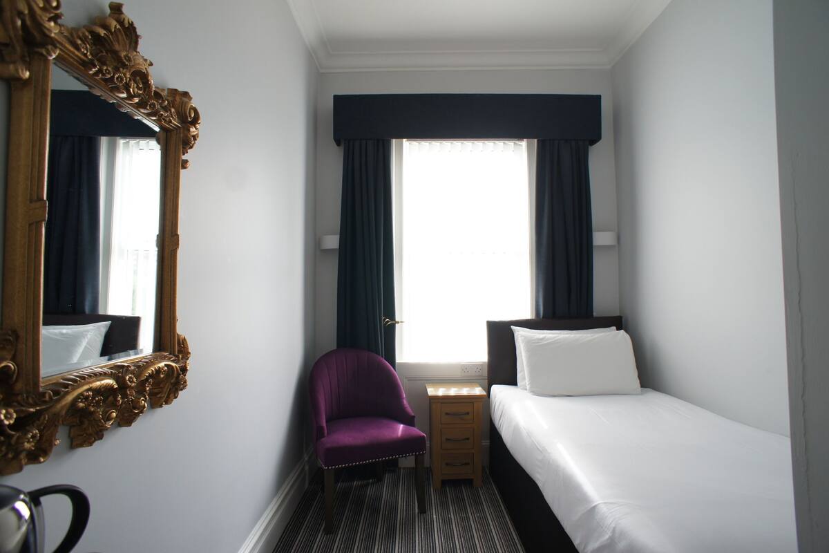 A hotel room with a bed, a chair, a nightstand, a mirror, and a window with curtains.