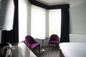 A hotel room with a bed, two purple armchairs, a small round table, and a coffee maker.