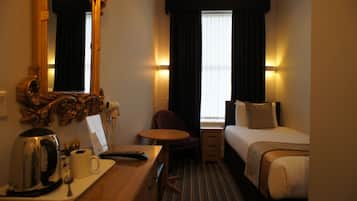 A hotel room with a bed, a desk with a kettle, a chair, a small table, and a mirror.