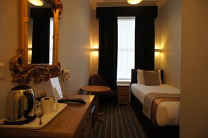A hotel room with a bed, a desk with a kettle, a chair, a small table, and a mirror.