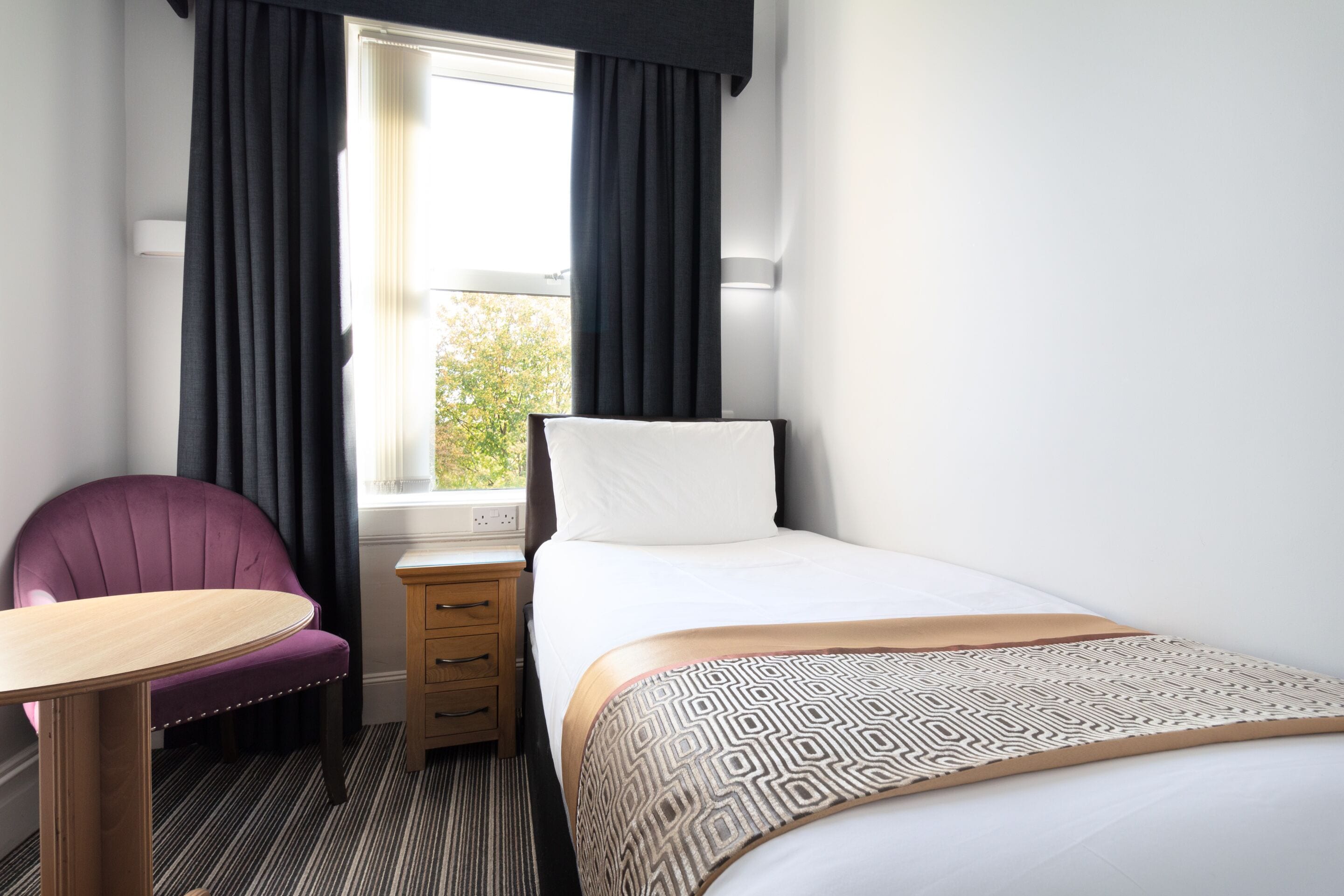 A hotel room with a bed, a chair, a small table, and a window with curtains.