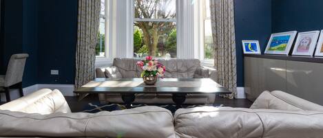 Una sala de estar con un sofá color beige, una mesa de centro con un jarrón de flores y vistas al exterior a través de un ventanal grande.
