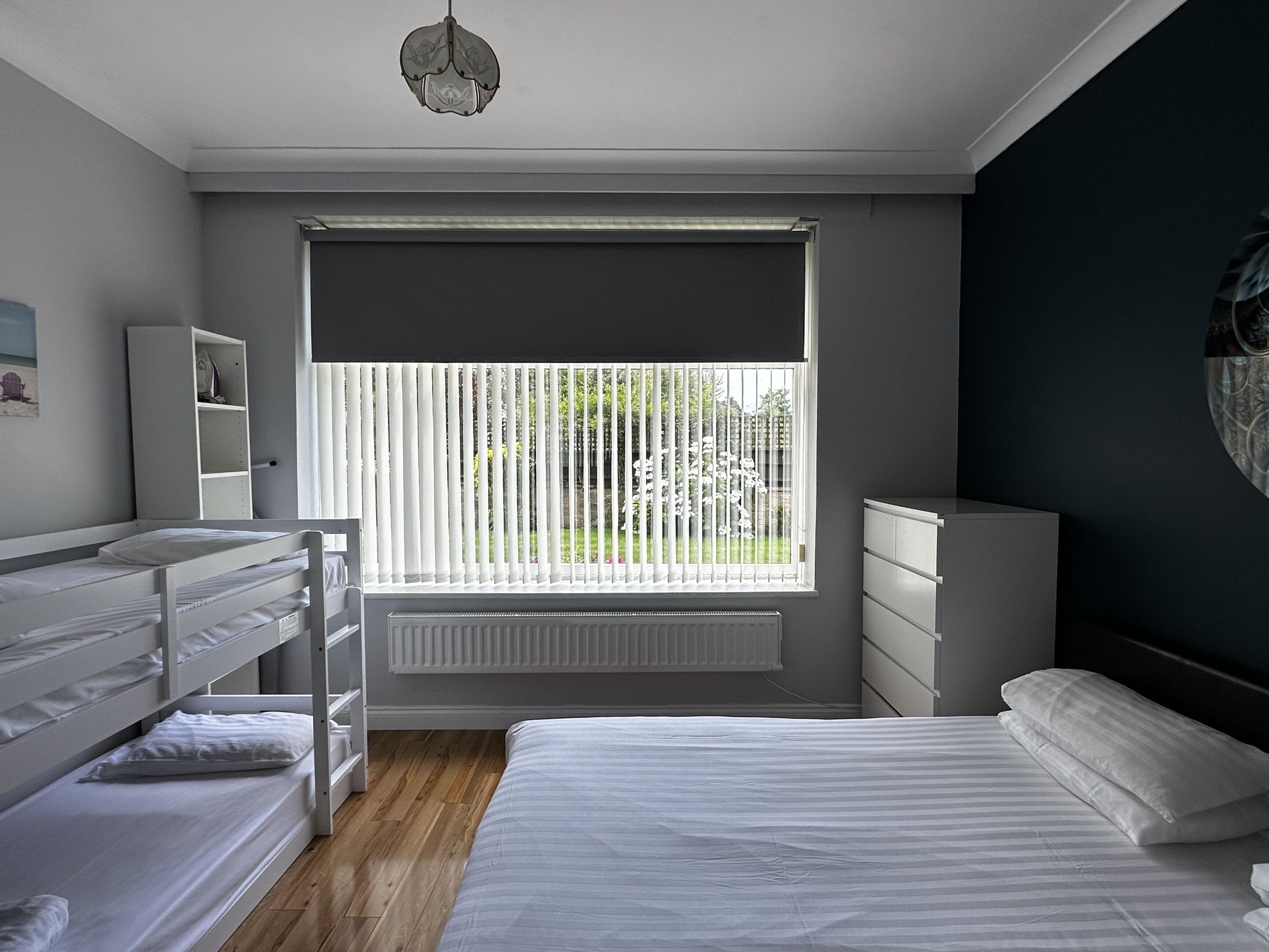 A bedroom with a bunk bed, a single bed, a radiator, a window with blinds, a wardrobe, and a hanging light fixture.