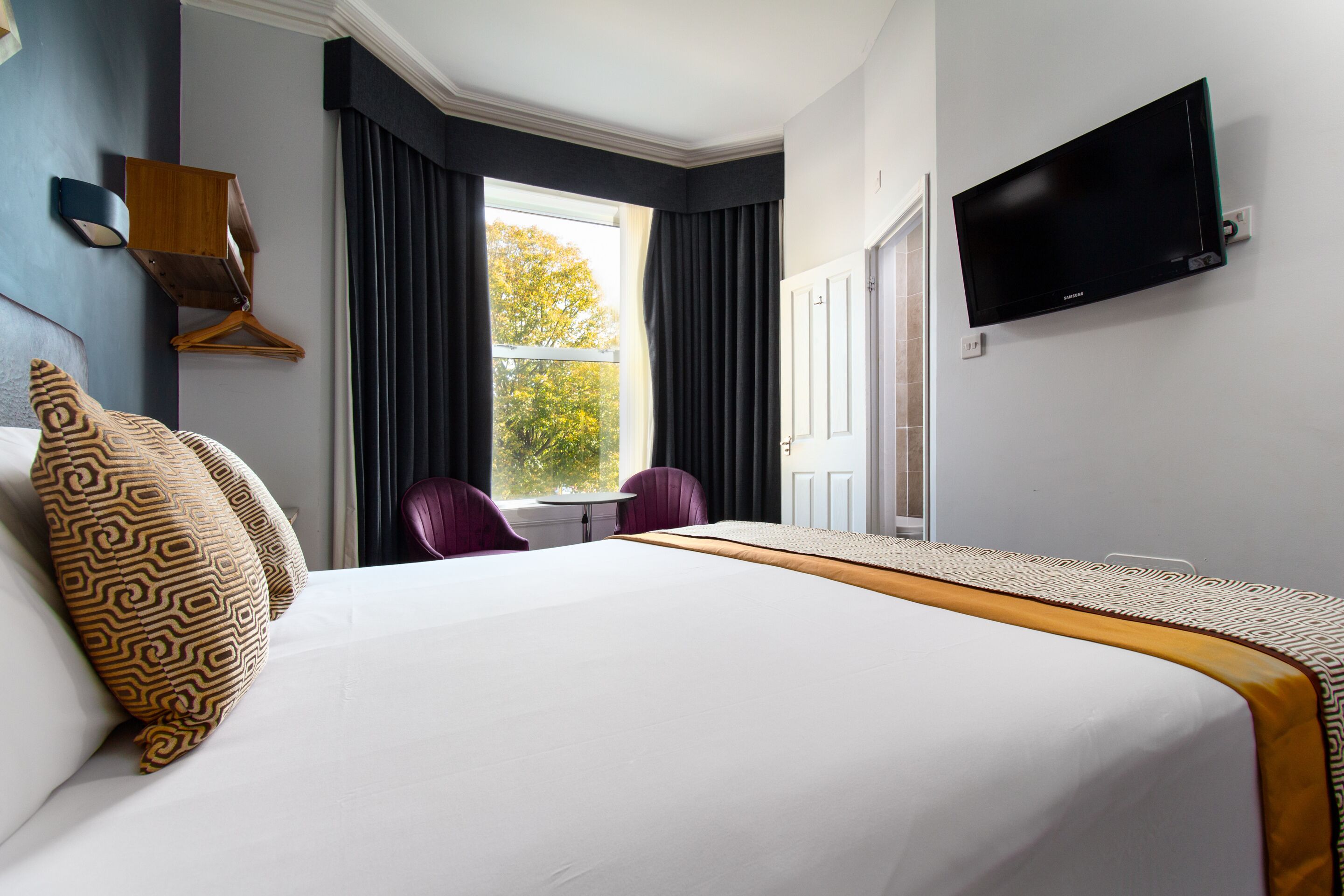 Een hotelkamer met een groot bed, een aan de muur bevestigde televisie, een raam met gordijnen en een stoel.