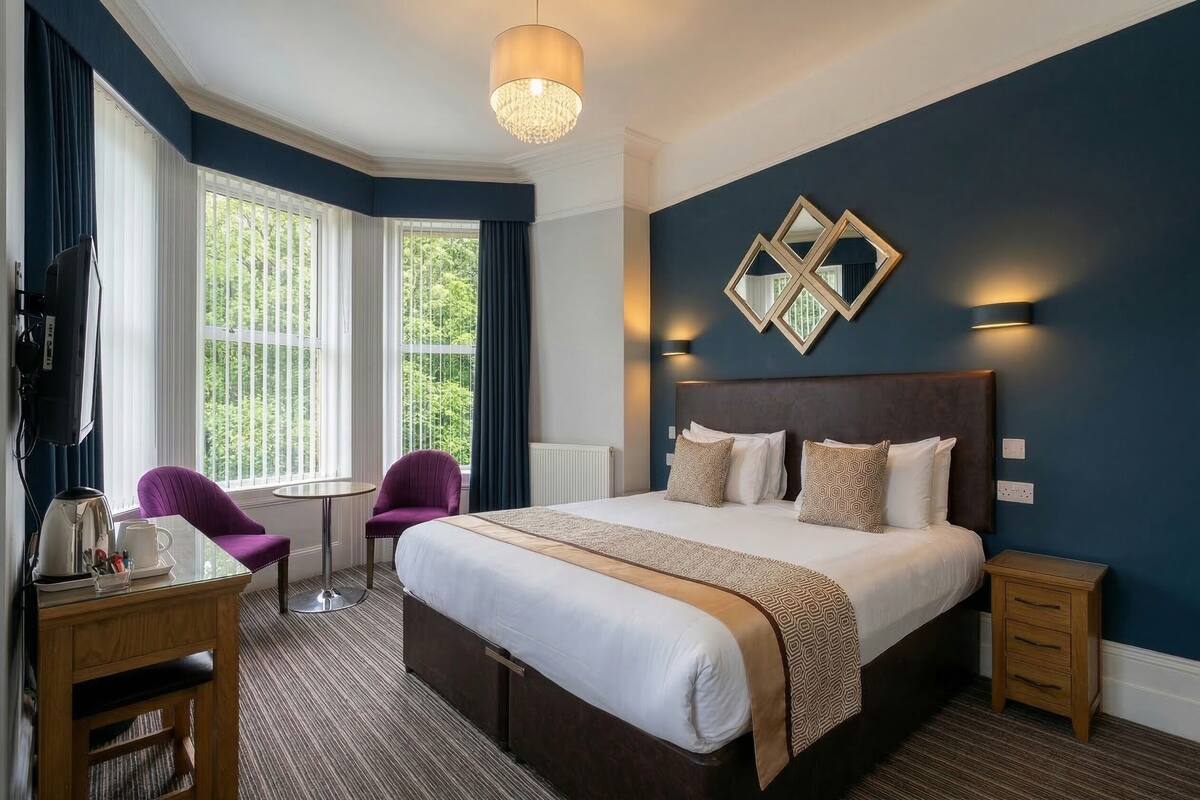 A hotel room with a large bed, a small table with a chair, a window with curtains, and a wall-mounted light fixture.