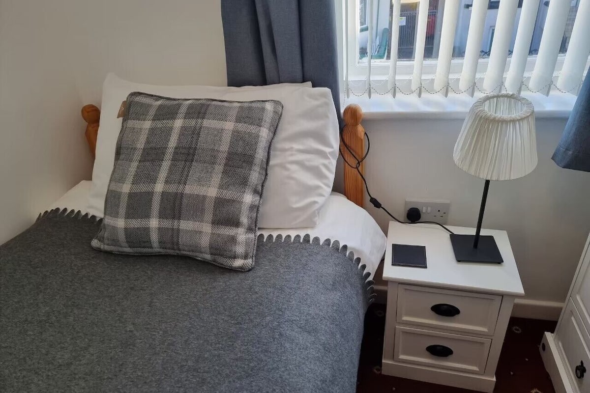A neatly made bed with a grey blanket and a plaid pillow, a bedside table with a lamp, and a window with blinds.