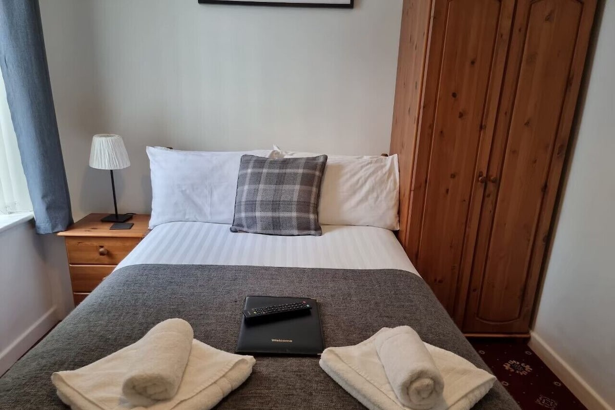 A neatly made bed with a plaid pillow, a remote control, and towels on a gray blanket.