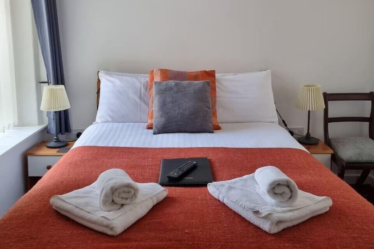 A hotel room with a bed, a painting of a beach scene, a window with curtains, a bedside table with a lamp, and a chair.
