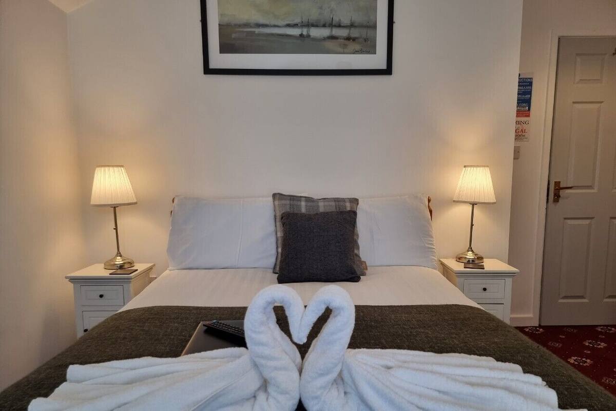 A hotel room with a bed, two bedside tables with lamps, a painting on the wall, and a heart-shaped towel arrangement on the bed.