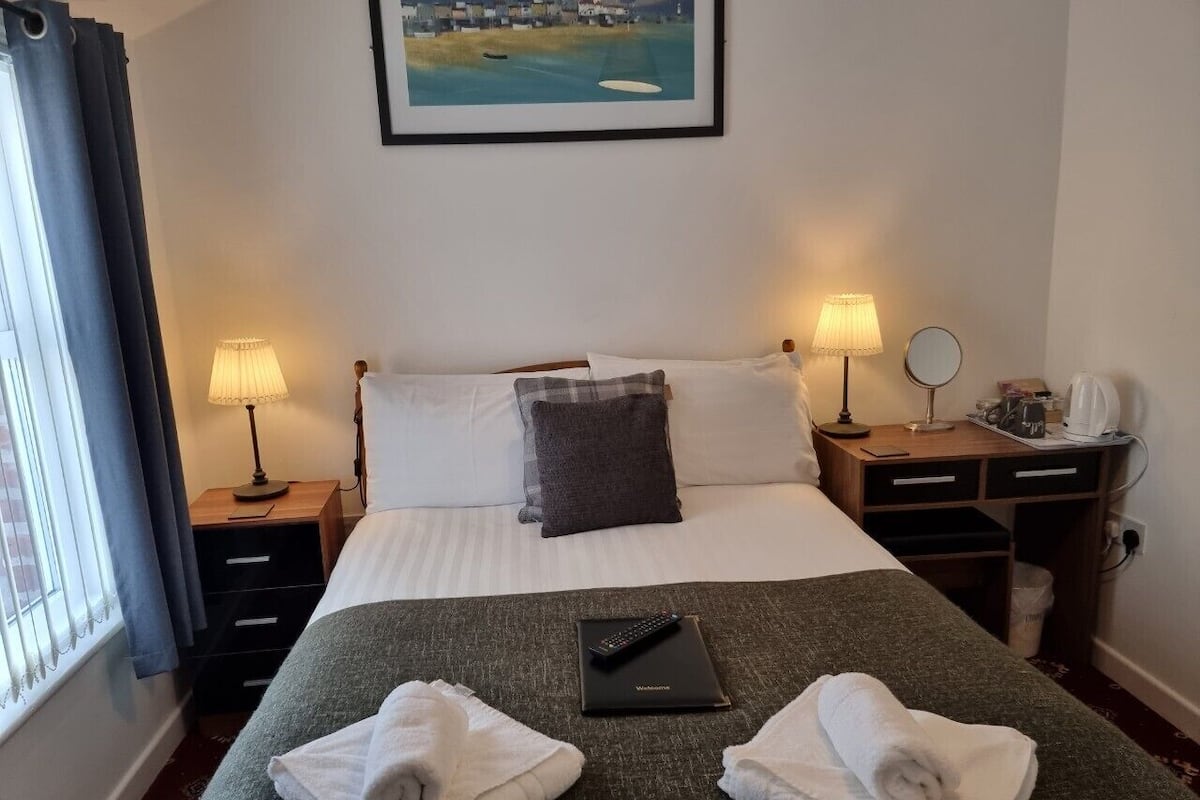 A hotel room with a bed, two bedside lamps, a bedside table with a lamp, a small table with a mirror, a laptop, and two towels on the bed.